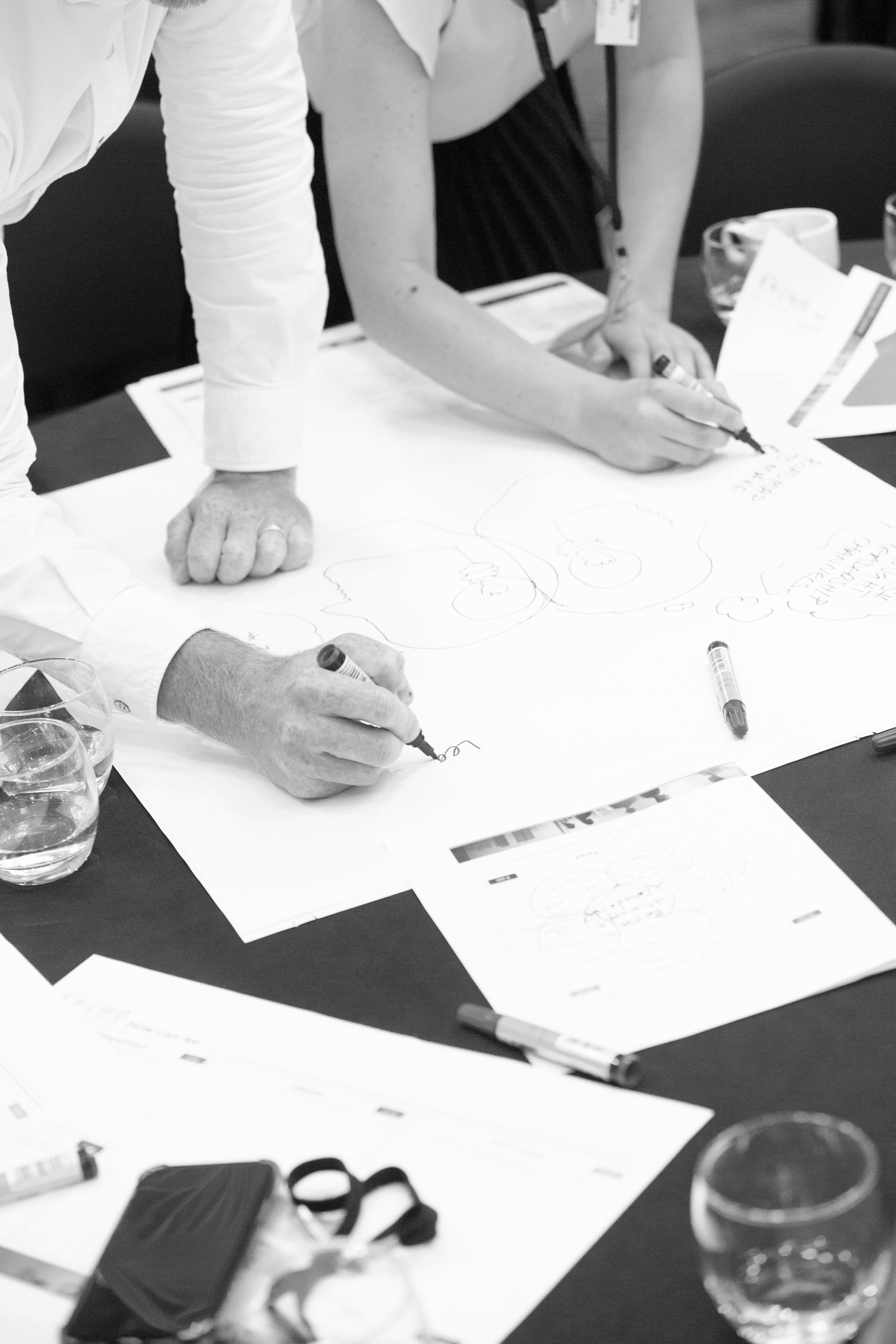 The arms of two people writing on a large piece of white paper during a workshop