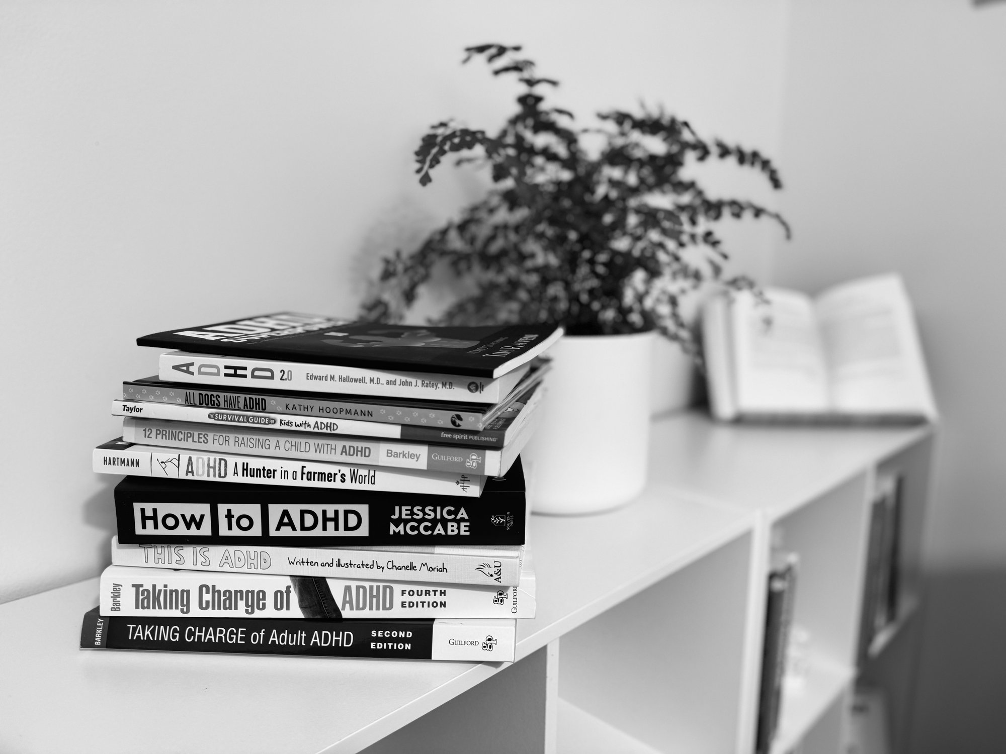 A pile of books about ADHD on a white shelf, with a plant, and open book in the background