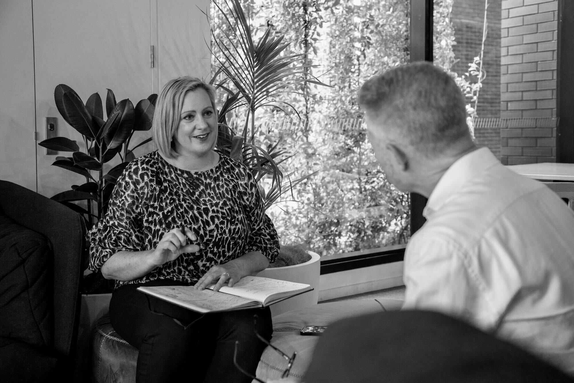 Jenny Turner, seated, with a notebook during a coaching session, talking to a man in a white shirt, who looks on.