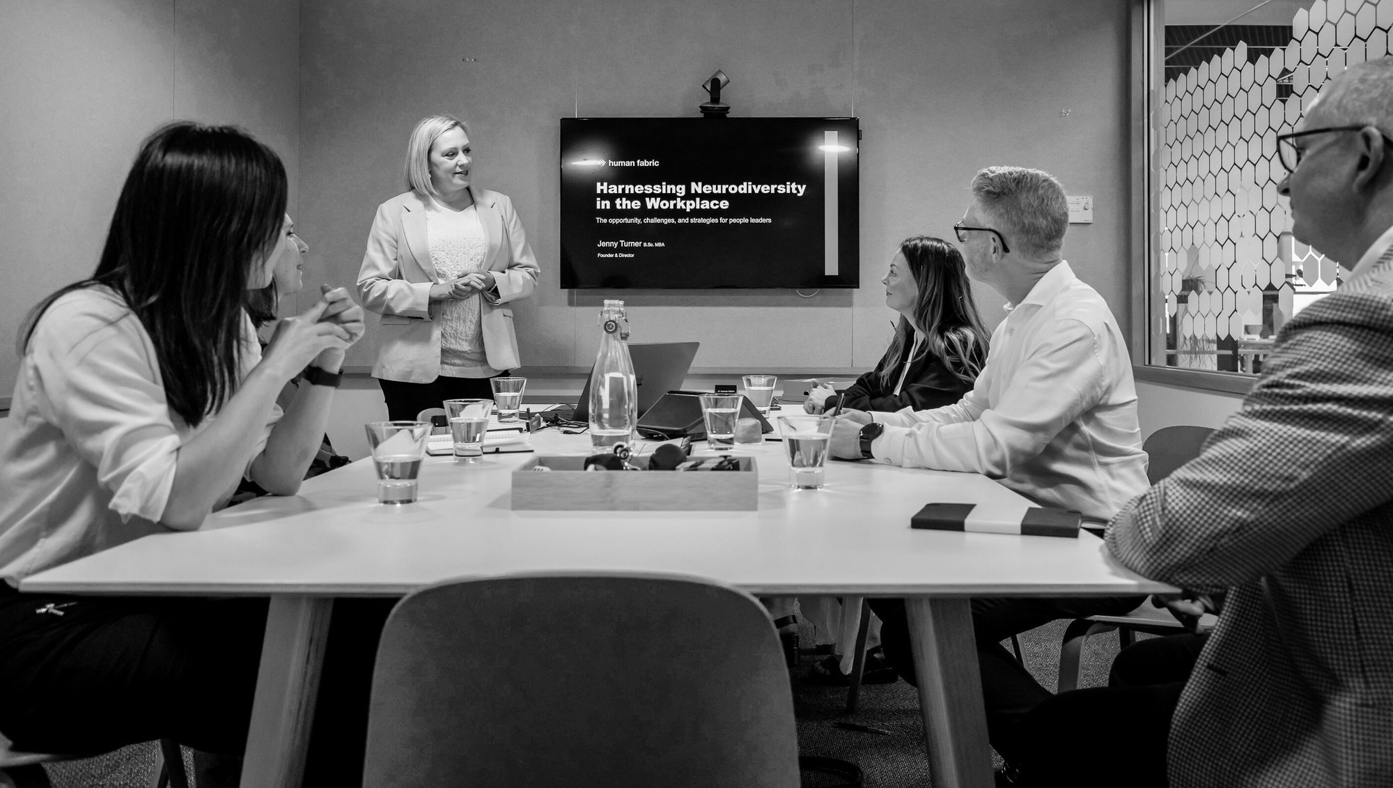 5 people in a room listening to Jenny talk, with a presentation on Harnessing Neurodiversity in the Workplace on the screen