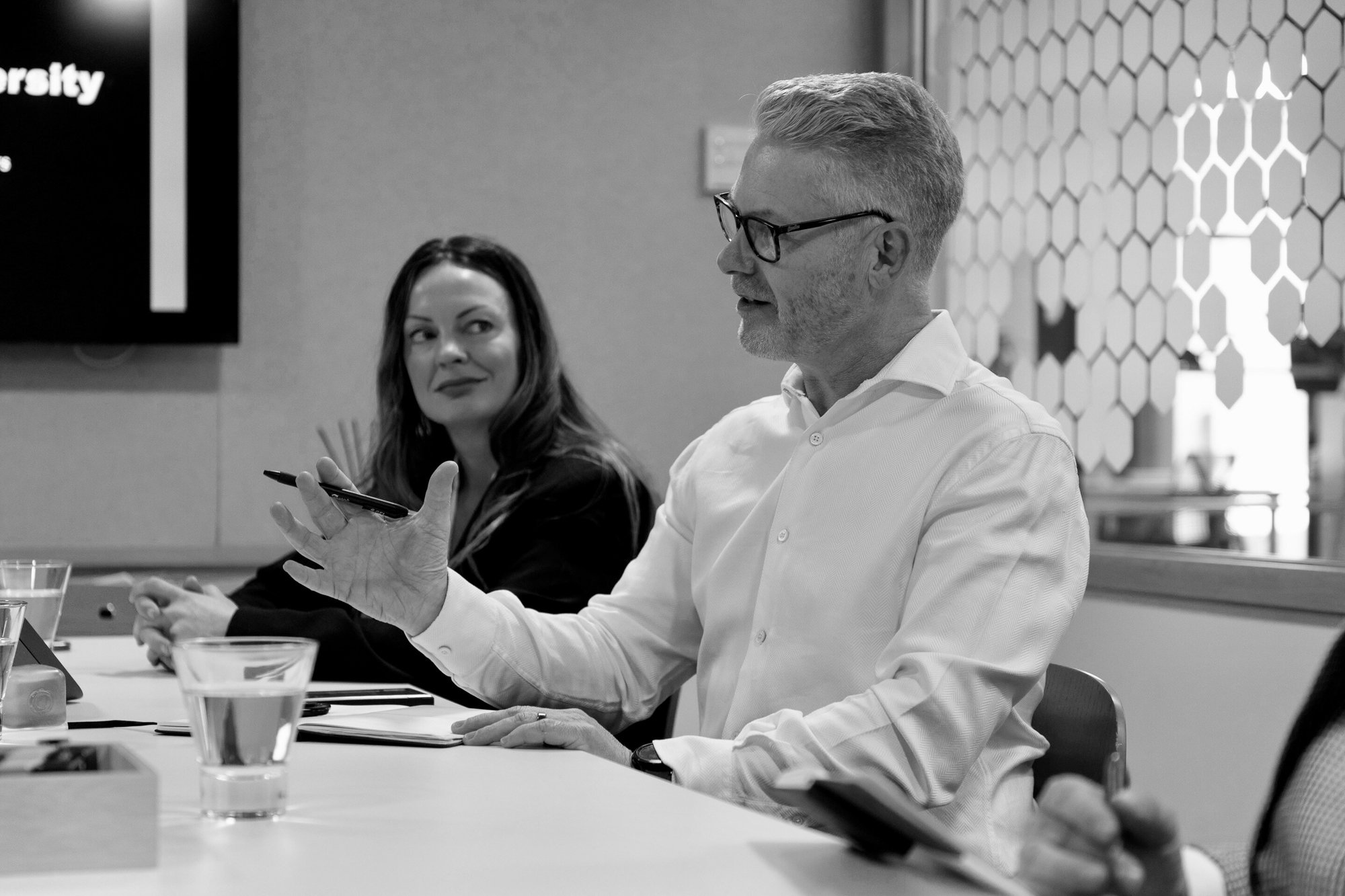 A man in a white shirt with his hand up, talking to the presenter who is out of view, while another woman looks on, seated at a table. 