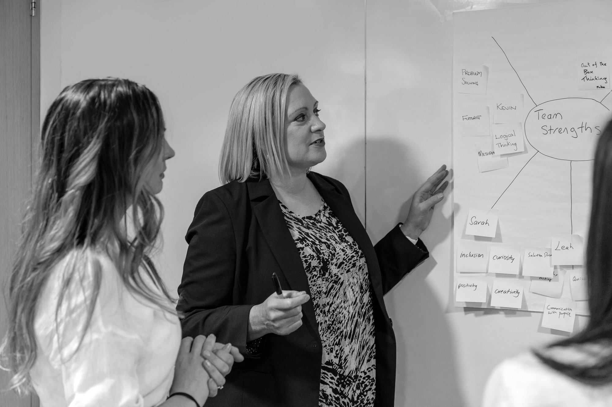 Jenny and a workshop participant standing at a wall and talking to a piece of paper with post it notes on it 