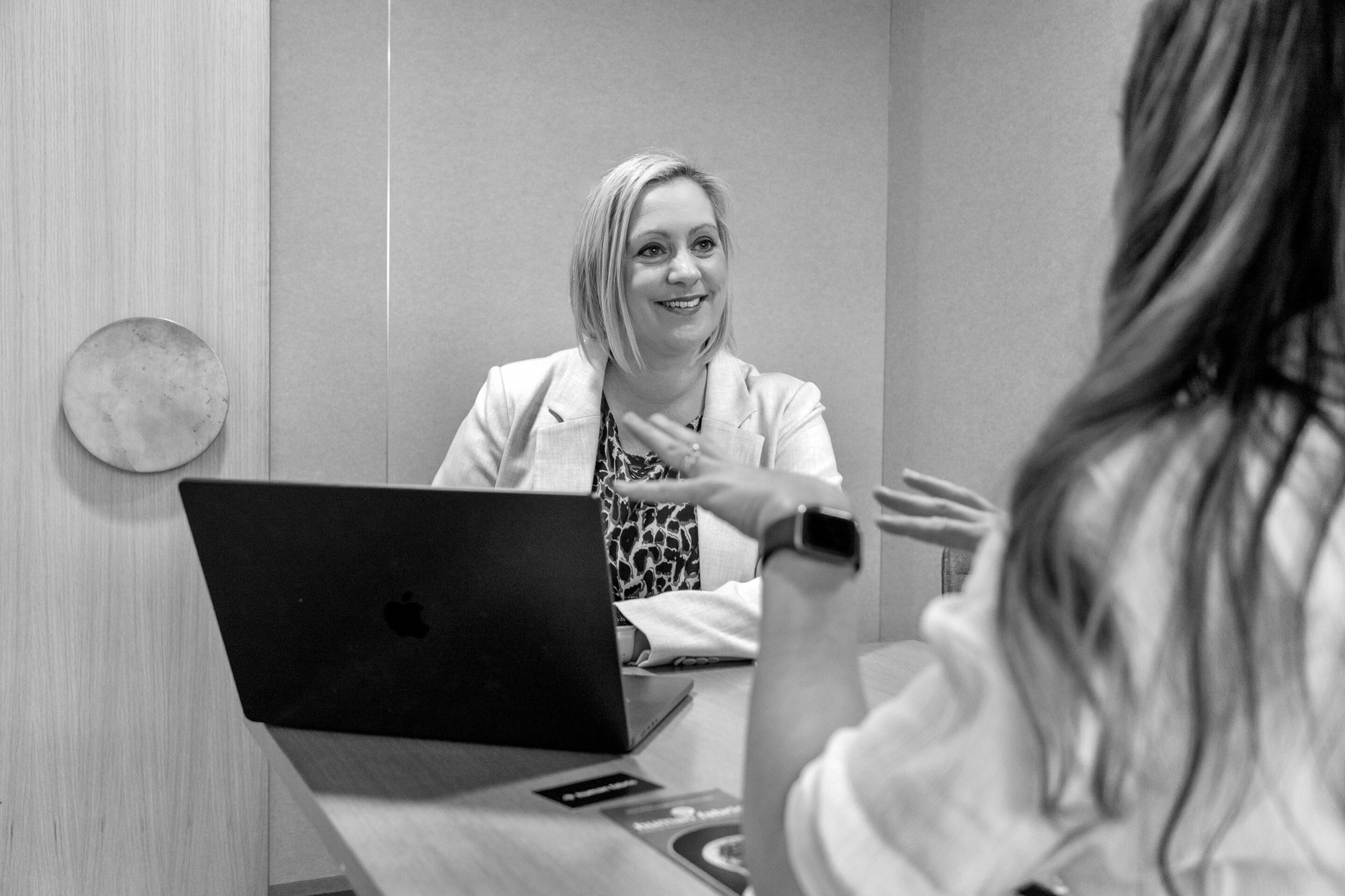 Jenny sitting in front of her laptop, while meeting with a woman whose face we can't see, and smiling during the conversation