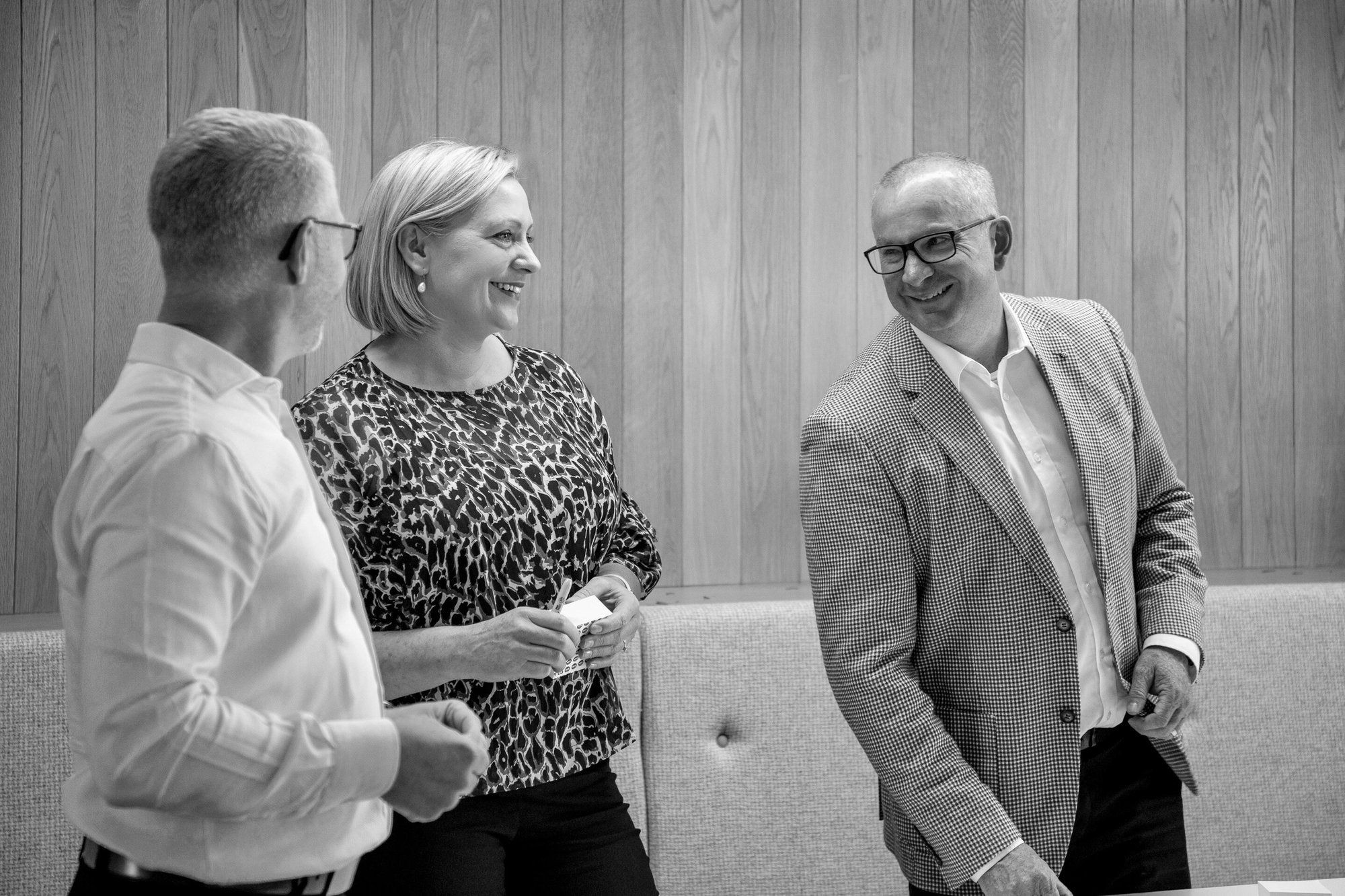 Three people in office attire laughing and talking - a woman and two men.