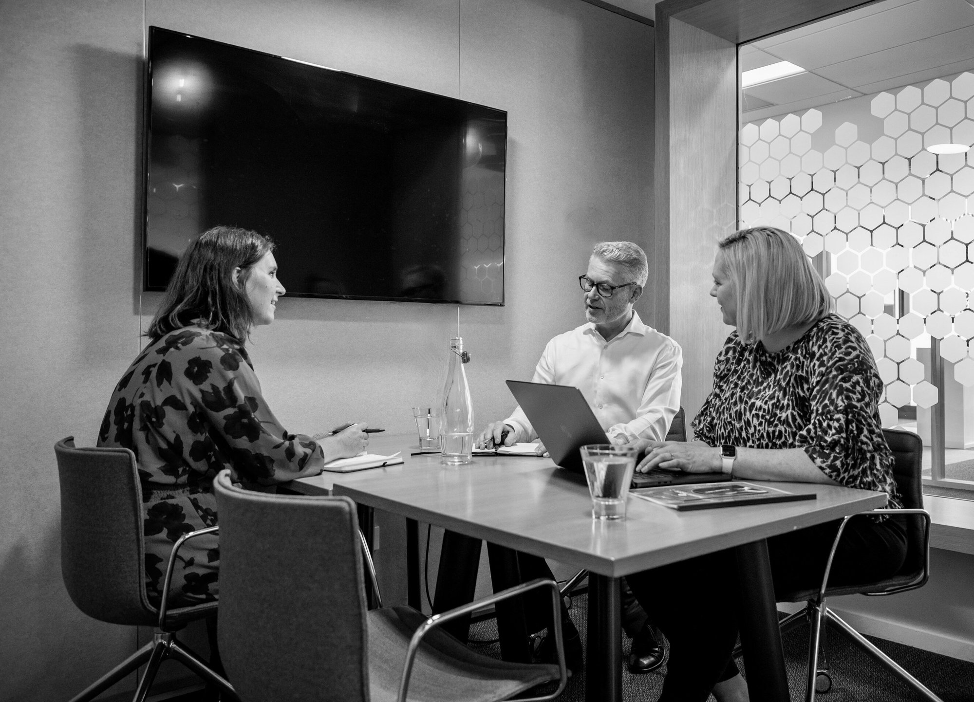 Two women and a man sitting at a table in a meeting room, having a discussion, one with a laptop out.