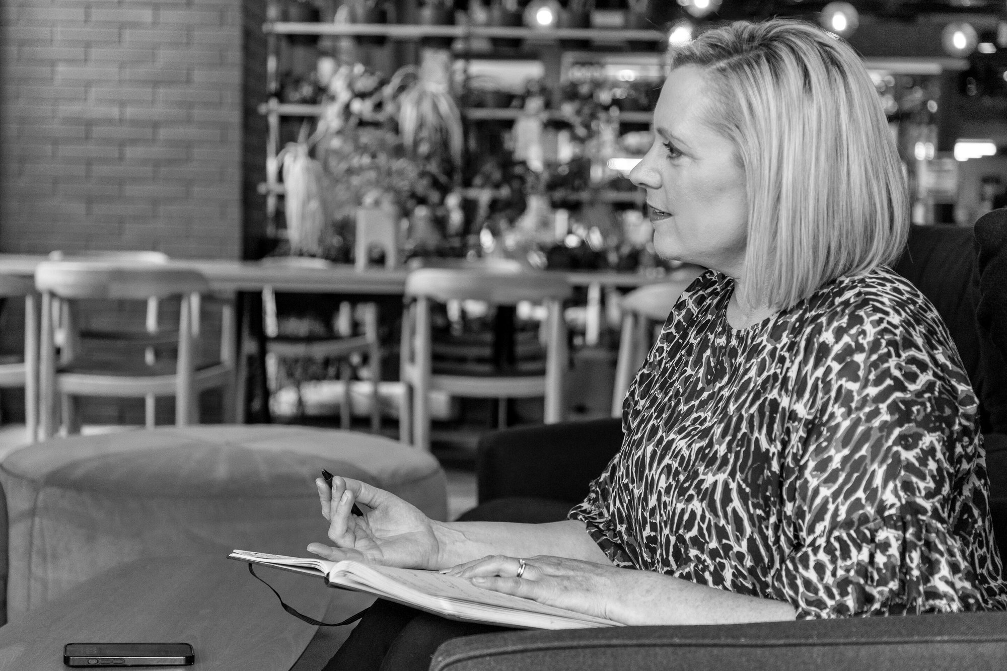 Jenny Turner talking to someone out of the picture, while seated, with a notebook and pen in hand.