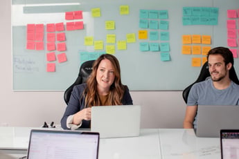 A male and a female in a meeting with others, laptops on the table, with sticky notes on a whiteboard behind them.
