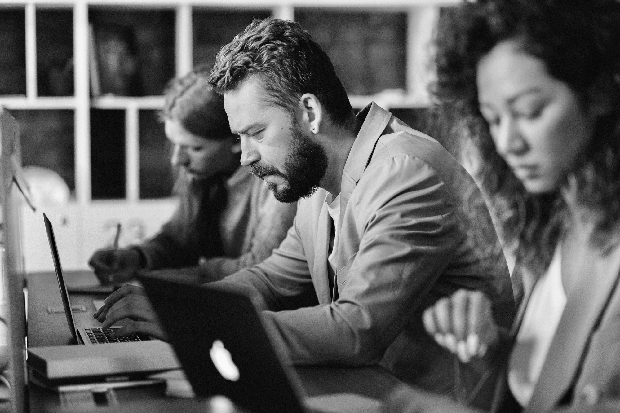 Three people working in a row along a work table, on their laptops. Focus is on a man in the middle who is looking thoughtful.